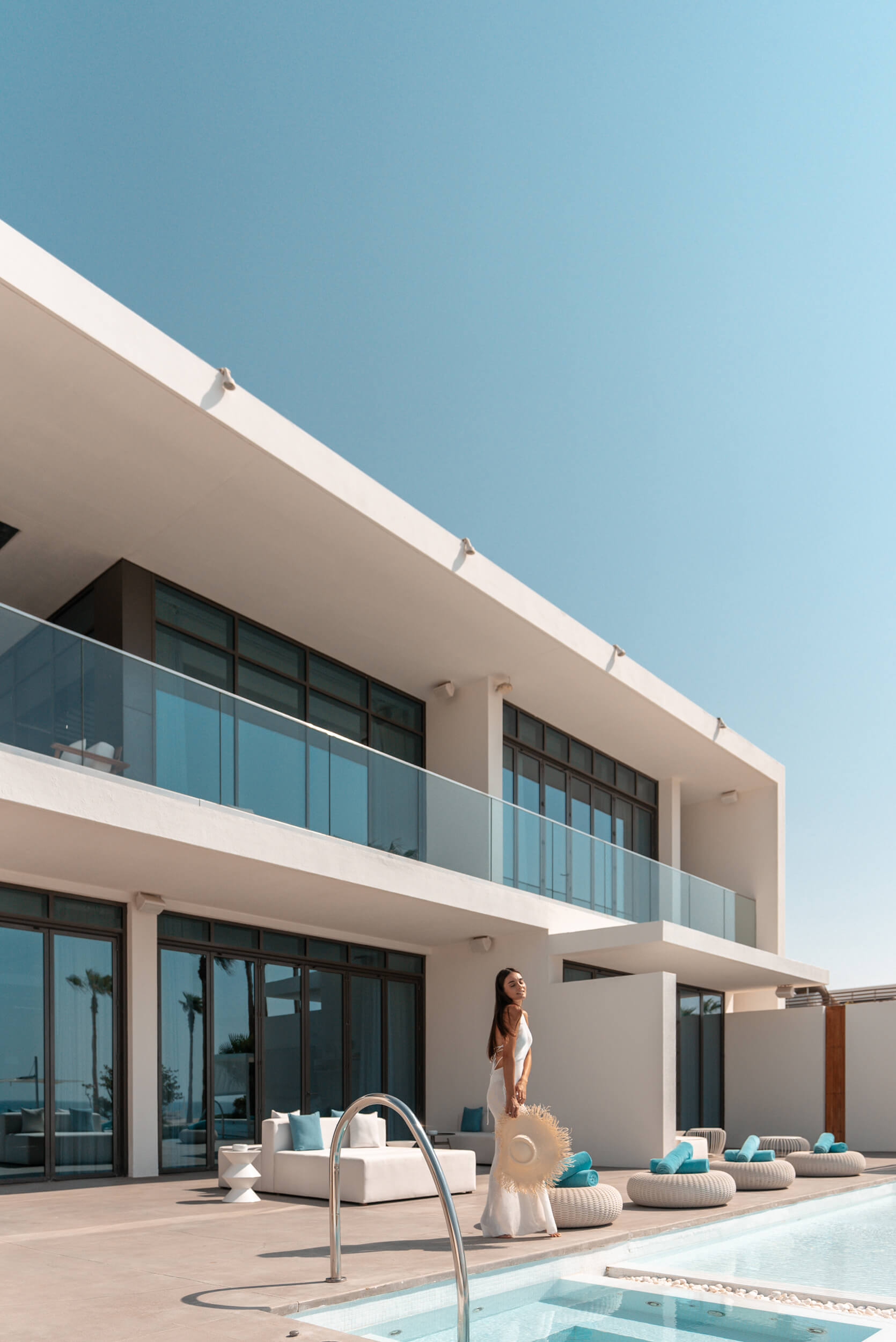 A women standing next to the Ultimate Villas pool, holding a straw hat. Sun Loungers and cushions surround the edges of the pool