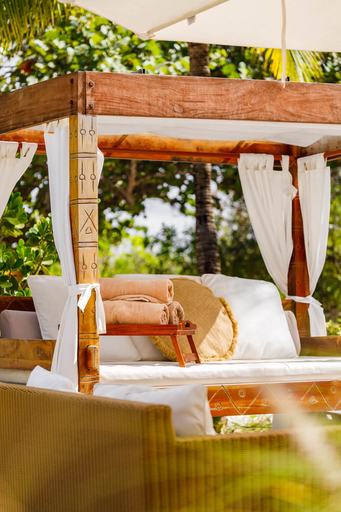 A Nikki Beach sun bed with carved wooden framings and white curtains tied back. The sun bed has multiple cushions and a small table with beach towels
